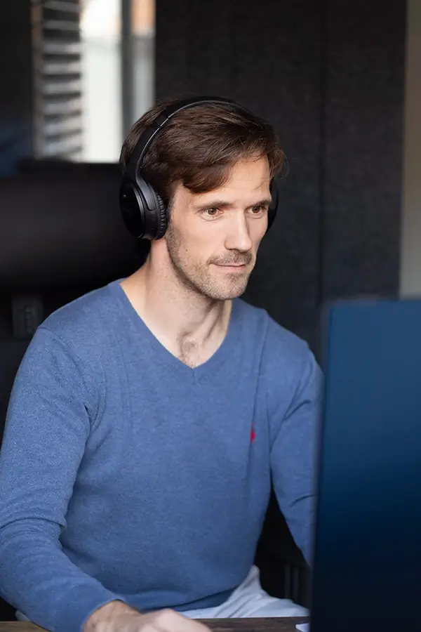 Stephen Spencer Voice Actor wearing a blue jumper and dark headphones. He's working on his computer, looking at the screen, sat inside.