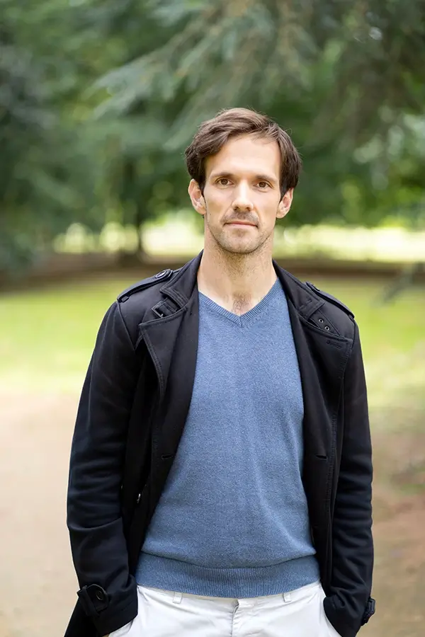 Stephen Spencer Voice Actor smiling, standing, with his hands in his pockets. He's outside in a park, wearing a dark casual jacket, blue jumper and white slacks.